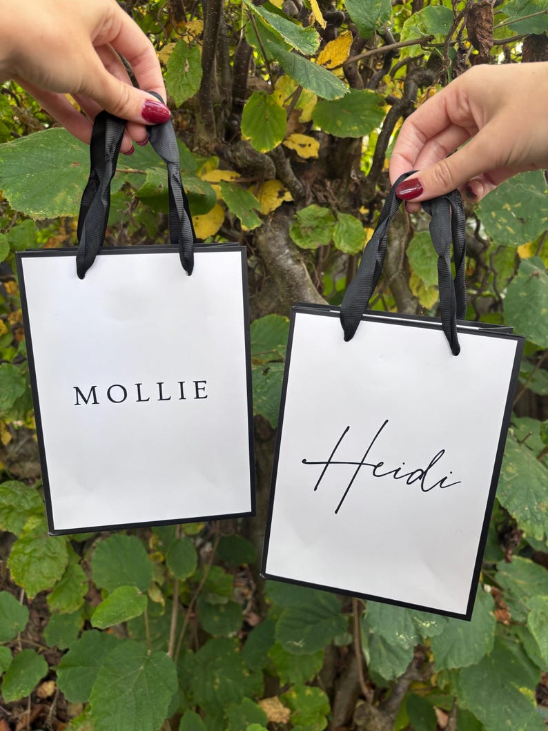 May include: Two white gift bags with black trim and black ribbon handles are held up against a backdrop of green and yellow leaves. One bag says "MOLLIE" and the other says "Heidi" in elegant black script.