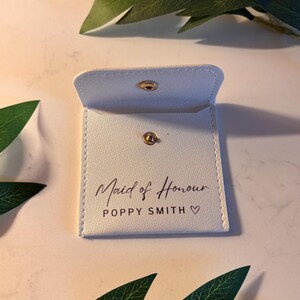 May include: A small, white pouch with a gold popper fastening. The words "Maid of Honour" and a name are printed on the front in a script font. The pouch is open. Green leaves are in the background.