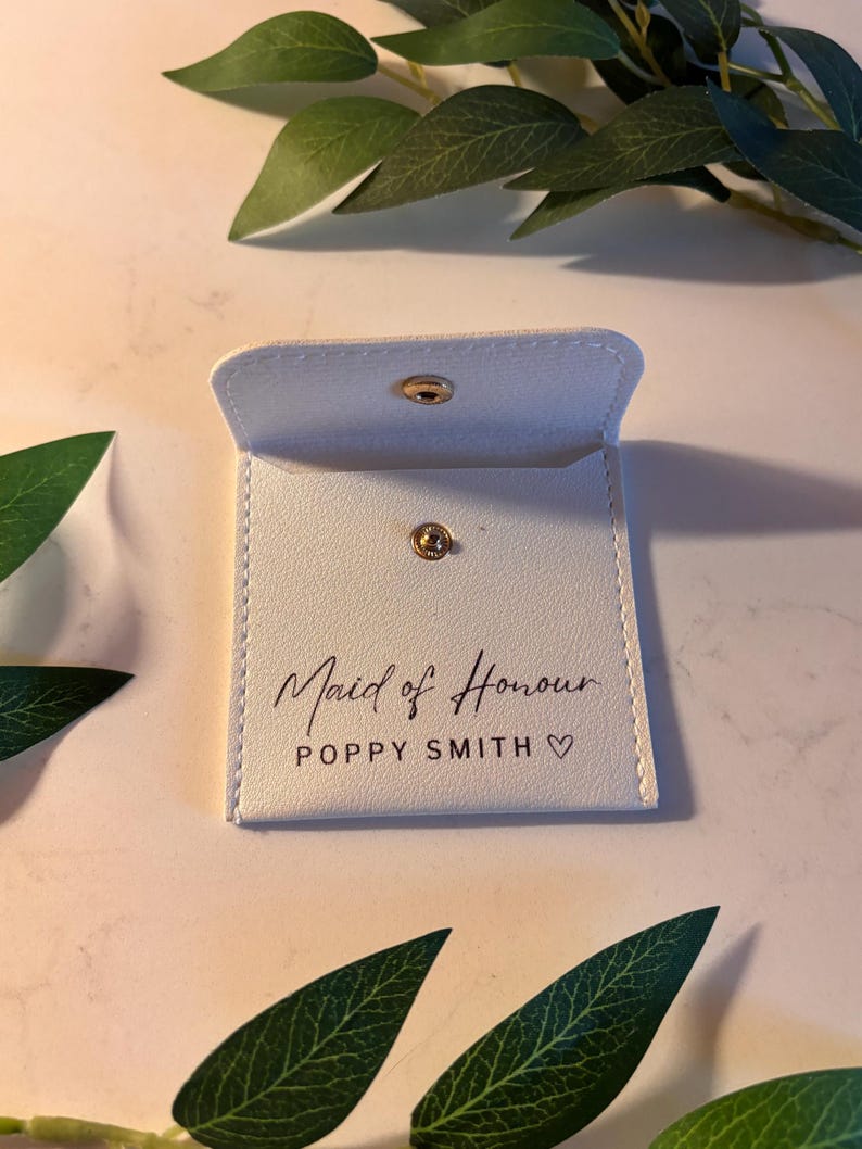May include: A small, white pouch with a gold popper fastening. The words "Maid of Honour" and a name are printed on the front in a script font. The pouch is open. Green leaves are in the background.
