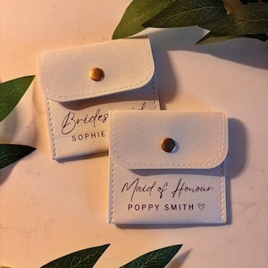 May include: Two small, off-white pouches with a flap closure and a gold-coloured snap. One pouch reads "Brides" and a name in script. The other reads "Maid of Honour" and a name, with a heart symbol. Green leaves are in the background.
