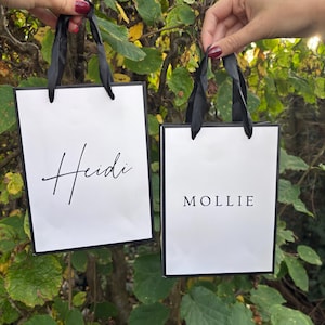 May include: Two white gift bags with black trim and black ribbon handles are held up against a backdrop of green foliage. One bag reads "Heidi" in cursive, the other reads "MOLLIE" in block letters.