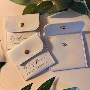 May include: Pale grey square pouches with gold press stud fastenings. One pouch reads "Bridesmaid" and another reads "Maid of Honour" with a name. Green leaves are in the background. These pouches are likely for small gifts.