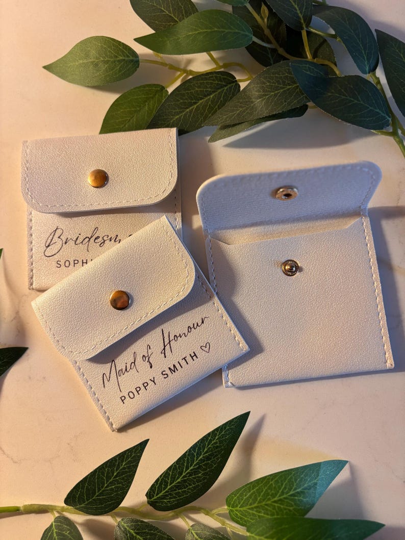 May include: Pale grey square pouches with gold press stud fastenings. One pouch reads "Bridesmaid" and another reads "Maid of Honour" with a name. Green leaves are in the background. These pouches are likely for small gifts.