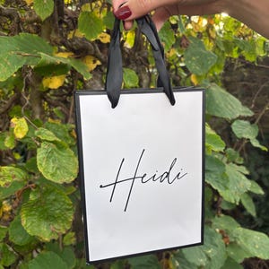 May include: A white gift bag with black trim and black ribbon handles. The name "Heidi" is written in elegant cursive script on the front of the bag. The bag is held up against a backdrop of green and yellow leaves.