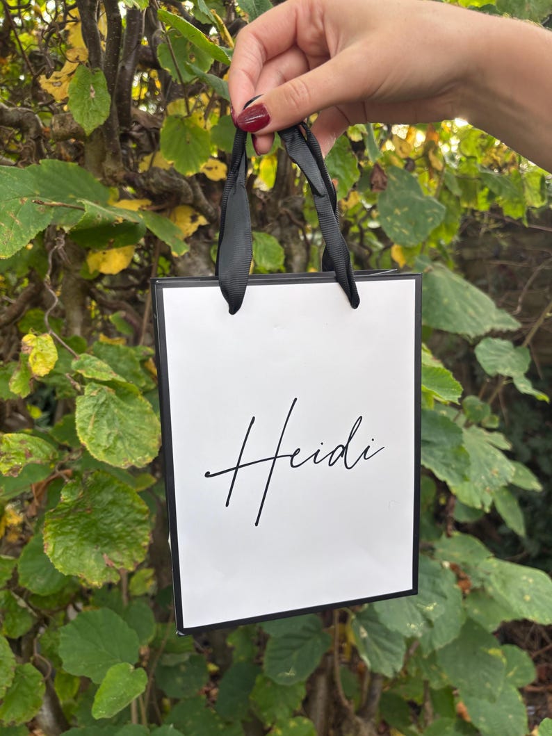 May include: A white gift bag with black trim and black ribbon handles. The name "Heidi" is written in elegant cursive script on the front of the bag. The bag is held up against a backdrop of green and yellow leaves.