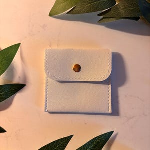 May include: A small, square, off-white pouch with a rounded flap and a gold-coloured button closure. The pouch is made of a smooth material and has white stitching around the edges. Green leaves are in the background.