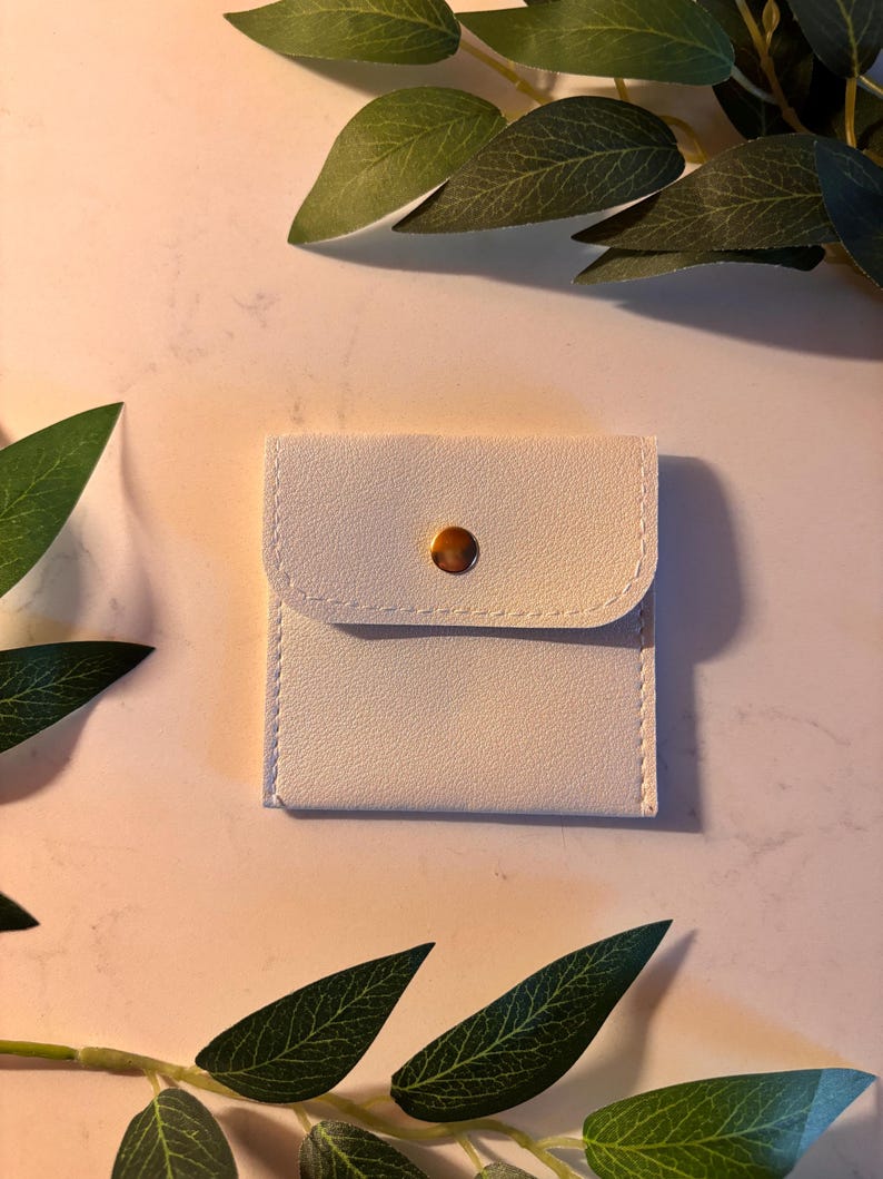 May include: A small, square, off-white pouch with a rounded flap and a gold-coloured button closure. The pouch is made of a smooth material and has white stitching around the edges. Green leaves are in the background.