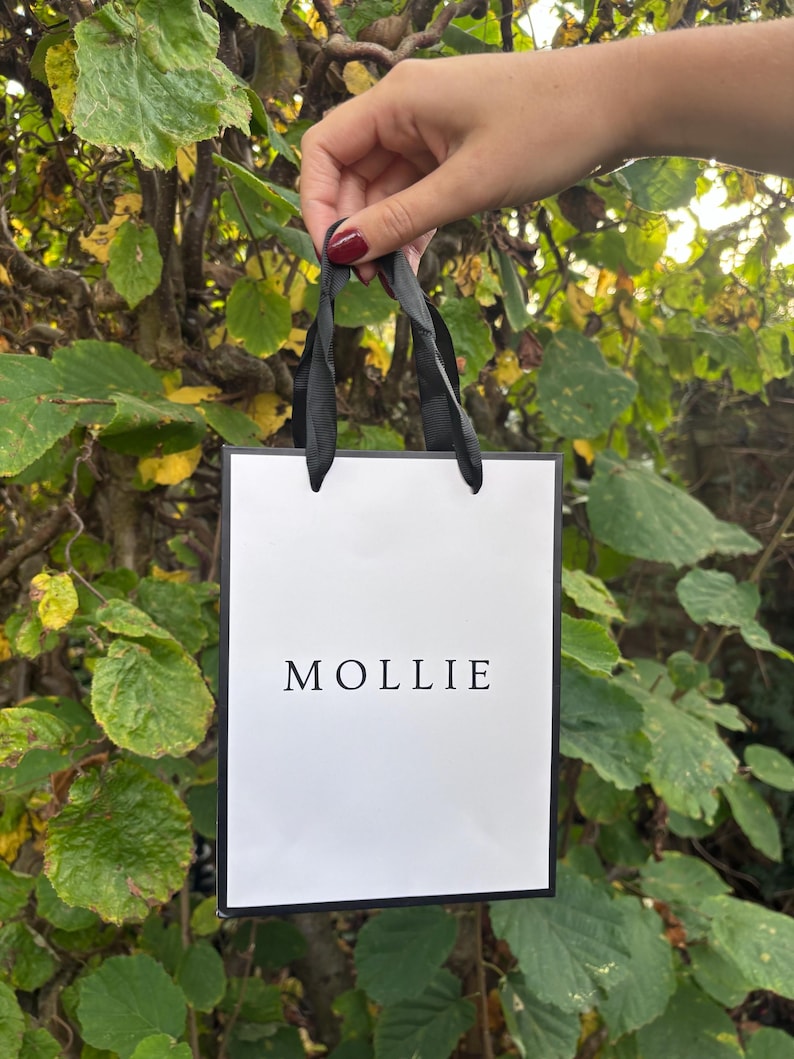 May include: A small white gift bag with the name "MOLLIE" printed in black. The bag has black trim and black ribbon handles. The bag is held up against a backdrop of green and yellow leaves.