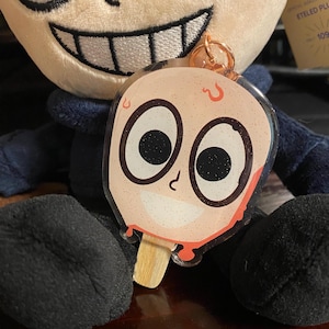May include: A clear plastic keychain charm shaped like a popsicle with a cartoon face. The face has large eyes with glitter, a small nose, and a smiling mouth. The popsicle is red and dripping with red sauce. The keychain is attached to a gold metal ring.