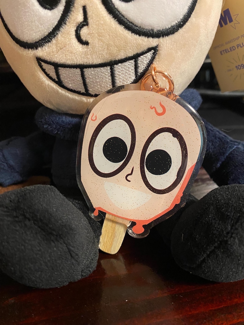 May include: A clear plastic keychain charm shaped like a popsicle with a cartoon face. The face has large eyes with glitter, a small nose, and a smiling mouth. The popsicle is red and dripping with red sauce. The keychain is attached to a gold metal ring.