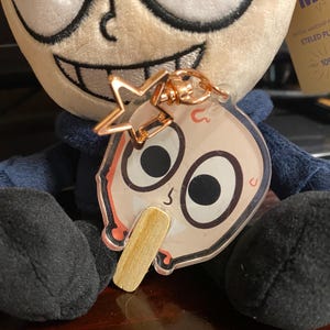 May include: A plush toy with a blue shirt and a large head with a wide smile. The toy is holding a clear keychain with a cartoon character on it. The keychain has a gold star charm attached to it.
