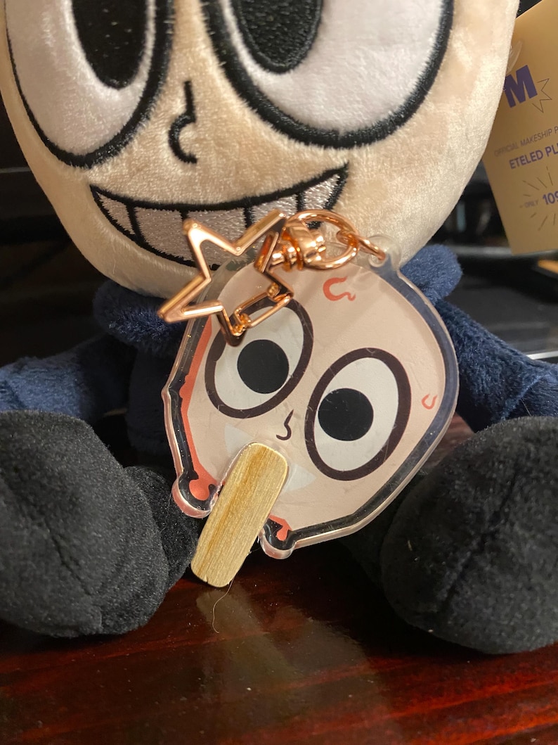May include: A plush toy with a blue shirt and a large head with a wide smile. The toy is holding a clear keychain with a cartoon character on it. The keychain has a gold star charm attached to it.