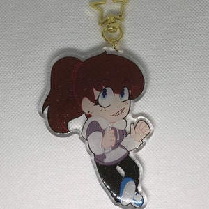 May include: A sparkly, clear acrylic keychain featuring a cartoon character with brown hair, a white and purple shirt, and blue shoes. The character is smiling and has a gold star charm attached to the top.