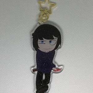 Op de afbeelding: Een zwarte en paarse glitter sleutelhanger met een cartoonfiguur met zwart haar, blauwe ogen en een rood mes. De sleutelhanger heeft een gouden stervormige hanger.
