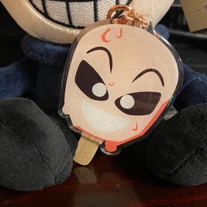 Può includere: Un peluche con un vestito nero e blu e un ampio sorriso, che tiene un portachiavi trasparente con un viso di cartone animato su un bastoncino di ghiacciolo. Il portachiavi ha glitter e una goccia rossa.