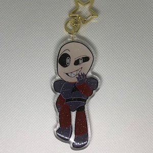 Puede incluir: Un llavero de acrílico transparente con purpurina que presenta un personaje de dibujos animados con una cara en blanco y negro, un cuerpo rojo y negro y un dije de estrella dorada.