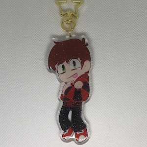 Pode incluir: Um chaveiro vermelho e preto com glitter, com um personagem de desenho animado com cabelo castanho, olhos verdes e uma camisa vermelha. O chaveiro tem um fecho em forma de estrela dourada.