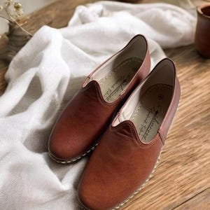 May include: A pair of brown leather slip-on shoes with a rounded toe and visible stitching along the sole. The interior lining is light-colored and features the text Charoog Handmade Footwear printed in black script on the heel area.