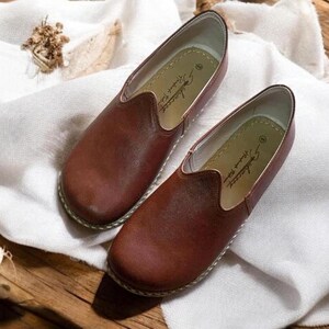 May include: A pair of brown leather slip-on shoes with a pointed toe and a simple design. The shoes have a white leather lining and a white leather sole. The brand name "Alessandro" is printed on the inside of the shoes.