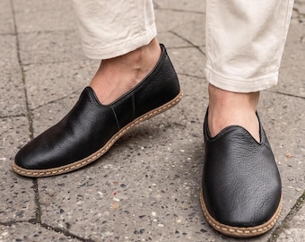 Puede incluir: Un par de zapatos sin cordones de cuero negro con un acabado de grano texturizado, punta redondeada y un detalle de costura en color tostado claro visible a lo largo de la suela. Los zapatos presentan una línea superior baja y un diseño sencillo.