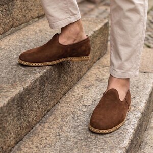 May include: A pair of dark brown suede slip-on shoes featuring a rounded toe, a low-cut collar, and a light tan stitched welt detail along the sole. The shoes are worn with light beige trousers, showcasing a simple and clean design profile.