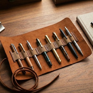 Può includere: Un astuccio in pelle marrone per penne con più penne di vari colori e design. L'astuccio è aperto, mostrando le penne fissate da cinturini in pelle. Un taccuino e un oggetto metallico sono visibili anche su una superficie di legno.