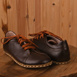 May include: A pair of brown leather shoes with orange laces. The shoes have a stitched seam around the sole and are sitting on a wooden surface.