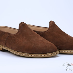 May include: A pair of brown suede slip-on shoes with a rounded toe, a high-cut vamp, and a tan sole featuring visible thick beige stitching around the perimeter. The logo for charooq Handmade Footwear is printed in the bottom right corner.