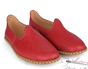 May include: A pair of red leather slip-on shoes with a brown stitched trim. The shoes are handmade and have a simple, elegant design.