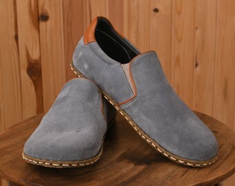 May include: A pair of gray suede slip-on shoes with brown leather accents and a stitched trim around the sole.