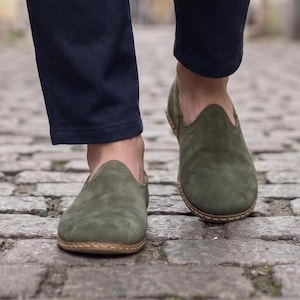 May include: A pair of olive green suede slip-on shoes with rounded toes and visible light-coloured stitching along the sole, worn with dark navy blue trousers while walking on a textured stone path.