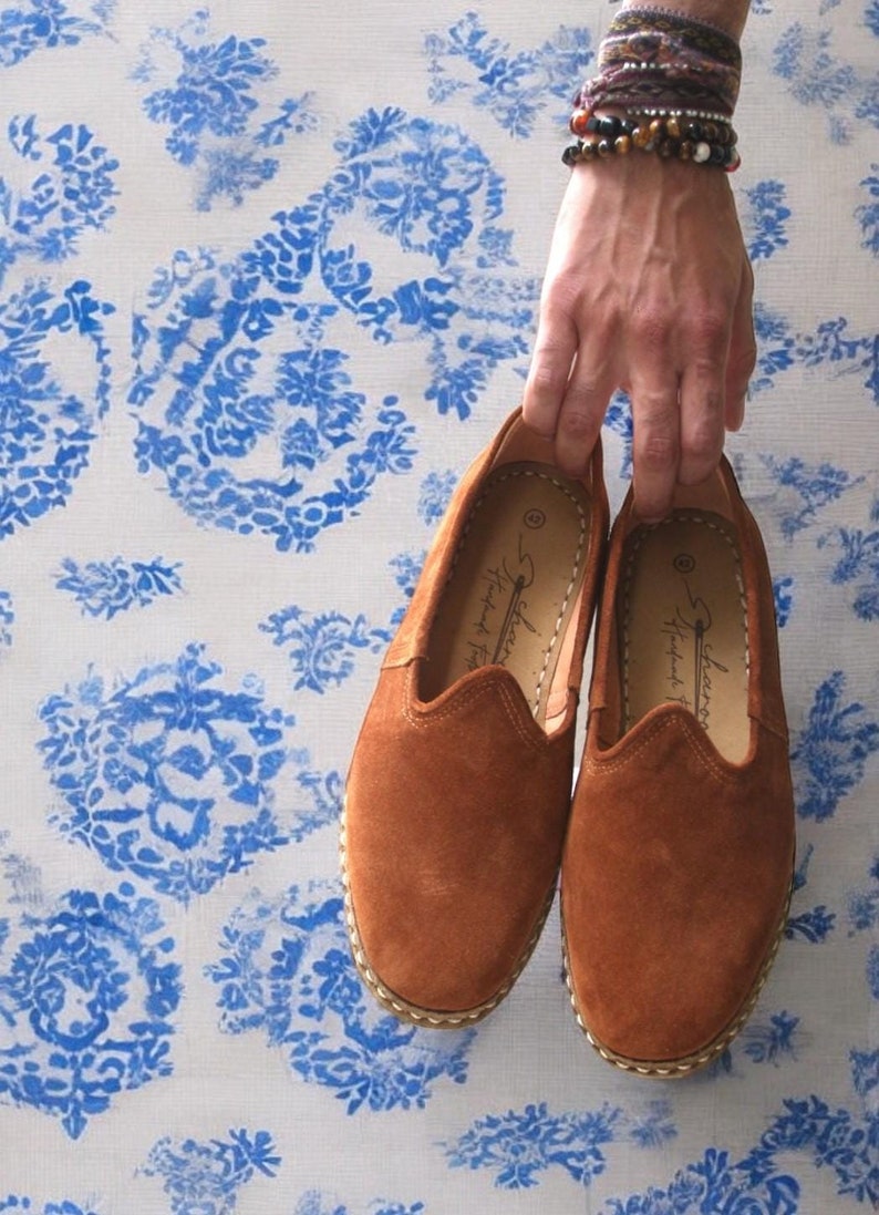 K&ouml;nnte beinhalten: Ein Paar braune Wildleder-Slipper mit runder Spitze und Ziern&auml;hten. Die Schuhe werden von einer Hand vor einem blau-wei&szlig; gebl&uuml;mten Hintergrund gehalten. Auf der Innensohle steht der Text "Scharang Handmade Footwear".