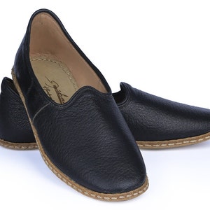 Op de afbeelding: Een paar zwarte leren slippers met een bruine gestikte rand. De slippers hebben een spitse neus en een platte zool.