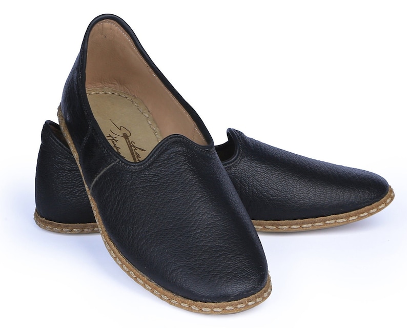 Op de afbeelding: Een paar zwarte leren slippers met een bruine gestikte rand. De slippers hebben een spitse neus en een platte zool.