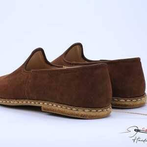 May include: A pair of brown suede slip-on shoes featuring a high-cut collar, a rounded toe, and a thick, light-colored stitched sole. A thin beige cord connects the two shoes. The text "charooq Handmade Footwear" appears in the bottom right.