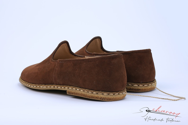 May include: A pair of brown suede slip-on shoes featuring a high-cut collar, a rounded toe, and a thick, light-colored stitched sole. A thin beige cord connects the two shoes. The text "charooq Handmade Footwear" appears in the bottom right.