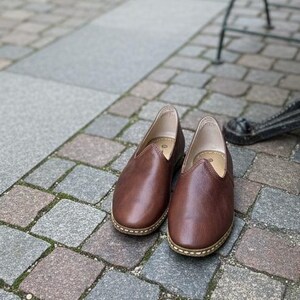 May include: A pair of brown leather slip-on shoes with a rounded toe and a stitched sole. The shoes are placed on a cobblestone surface, suggesting outdoor use. The shoes appear to be unworn.