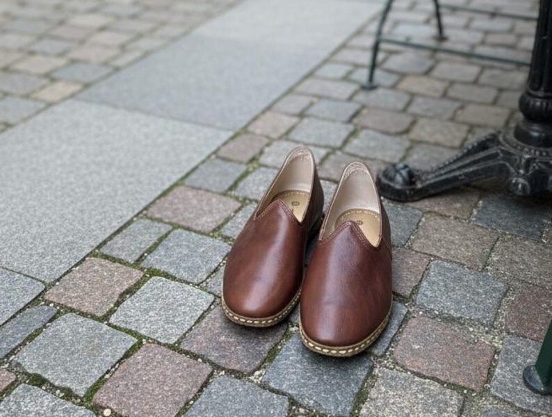 May include: A pair of brown leather slip-on shoes with a rounded toe and a stitched sole. The shoes are placed on a cobblestone surface, suggesting outdoor use. The shoes appear to be unworn.