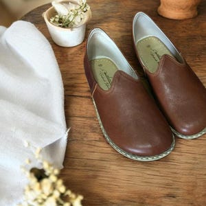 May include: A pair of brown leather slip-on shoes with white stitching around the edges. The shoes have a pointed toe and a flat sole. The brand name "Handmade for You" is visible on the inside of the shoes.