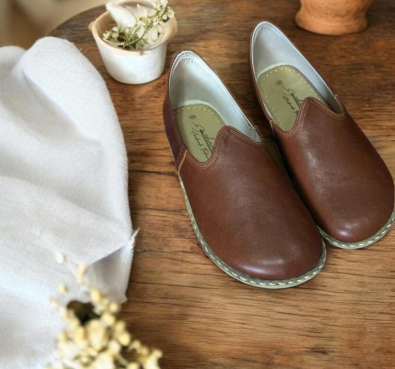May include: A pair of brown leather slip-on shoes with white stitching around the edges. The shoes have a pointed toe and a flat sole. The brand name "Handmade for You" is visible on the inside of the shoes.
