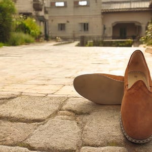 K&ouml;nnte beinhalten: Ein Paar braune Wildleder-Slipper. Ein Schuh steht aufrecht und zeigt die Oberseite und das Seitenprofil, w&auml;hrend der andere auf seiner Sohle liegt. Die Schuhe haben eine runde Spitze und eine gen&auml;hte Sohle.