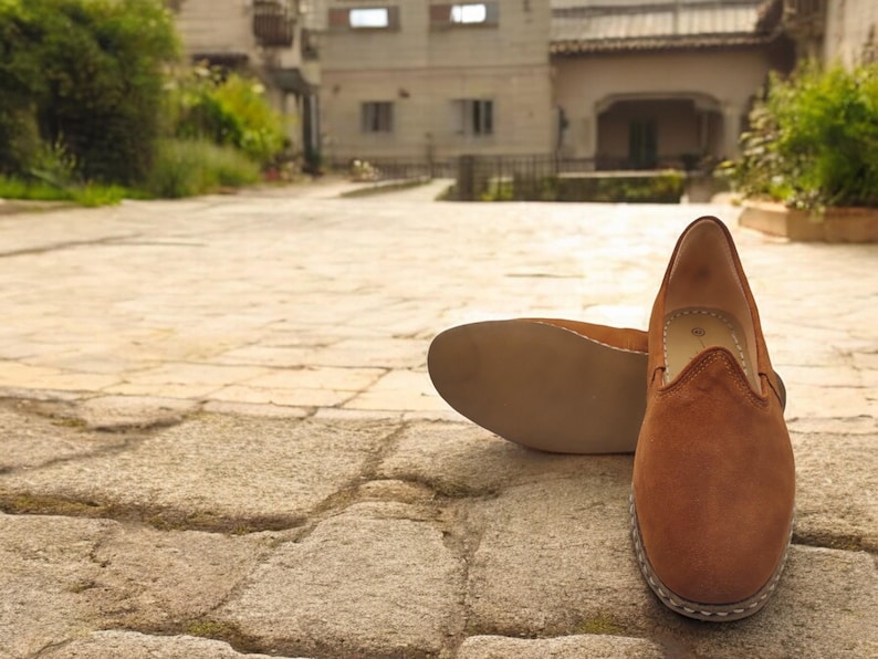 K&ouml;nnte beinhalten: Ein Paar braune Wildleder-Slipper. Ein Schuh steht aufrecht und zeigt die Oberseite und das Seitenprofil, w&auml;hrend der andere auf seiner Sohle liegt. Die Schuhe haben eine runde Spitze und eine gen&auml;hte Sohle.