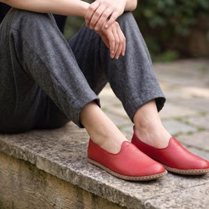 Puede incluir: Un par de zapatos sin cordones de cuero rojo con punta redondeada y suela marr&oacute;n claro. Los zapatos est&aacute;n sobre una superficie de piedra. La persona lleva pantalones grises.