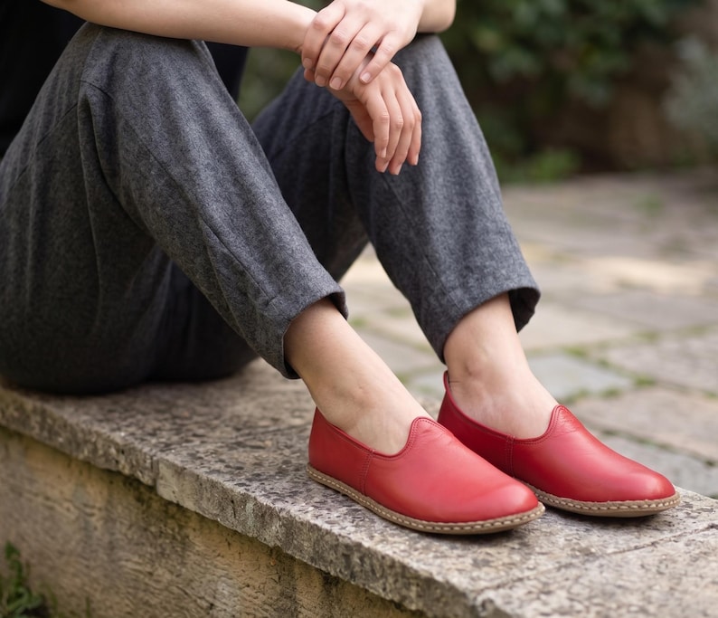 Puede incluir: Un par de zapatos sin cordones de cuero rojo con punta redondeada y suela marr&oacute;n claro. Los zapatos est&aacute;n sobre una superficie de piedra. La persona lleva pantalones grises.