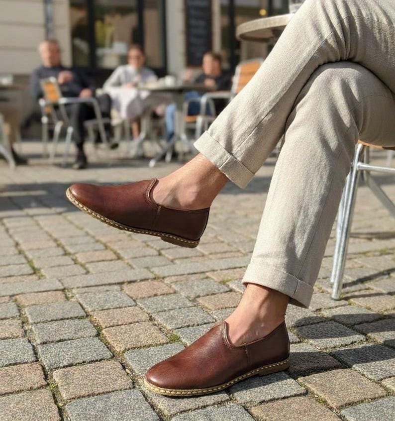 May include: Brown leather slip-on shoes with a rounded toe and a stitched sole. The shoes are worn with light-colored pants, suggesting a casual or semi-formal style. The image is taken outdoors on a cobblestone surface.