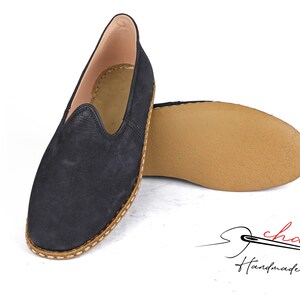 Puede incluir: Un par de zapatos negros de cuero con deslizamiento con una suela de cuero marr&oacute;n y un borde cosido. Los zapatos son hechos a mano y tienen un aspecto casual y c&oacute;modo.