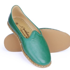 May include: A pair of green leather slip-on shoes with a brown sole and a white stitching detail around the edge. The shoes have a simple design and are made of soft leather.