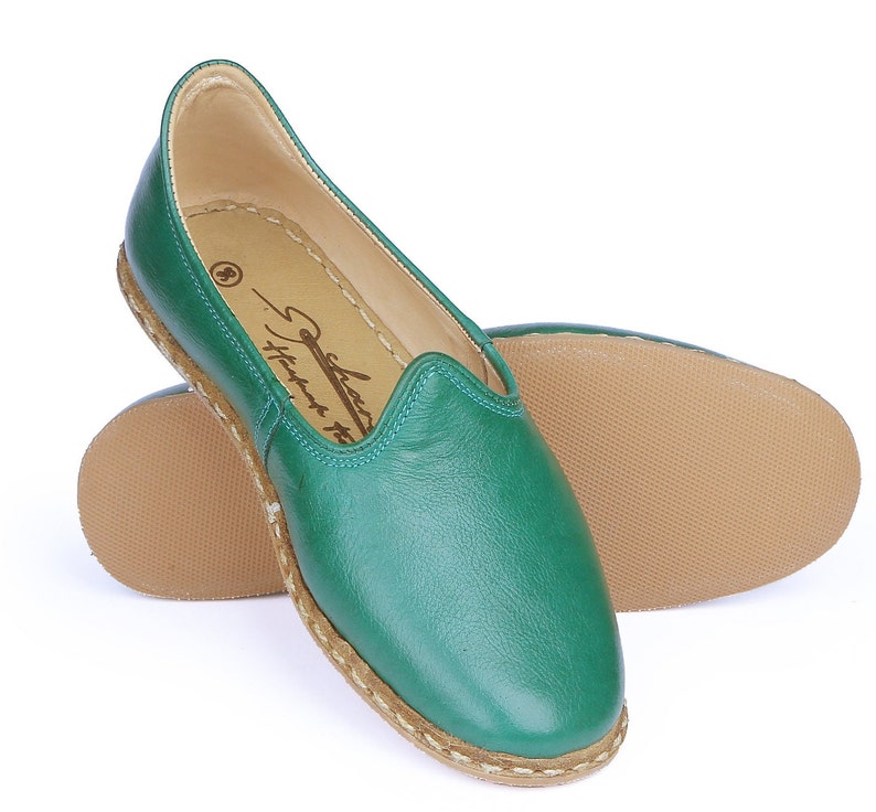 May include: A pair of green leather slip-on shoes with a brown sole and a white stitching detail around the edge. The shoes have a simple design and are made of soft leather.