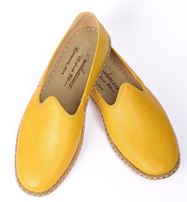 May include: A pair of yellow leather slip-on shoes with a stitched edge. The shoes have a simple design and are made of soft leather. The insole of the shoe has the text "Handmade with love, handcrafted shoes @charcoco.store".