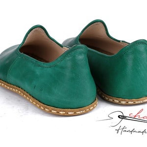 May include: A pair of green leather slip-on shoes with a brown stitched trim. The shoes are handmade and have a casual style.